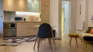 Una cocina moderna con microondas, horno y armarios de madera. Hay un comedor con una mesa redonda y una silla. El ambiente tiene piso de madera y un tapete con diseño geométrico.