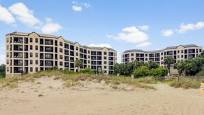 Un hotel frente a la playa con varios pisos, balcones y una playa de arena en primer plano.