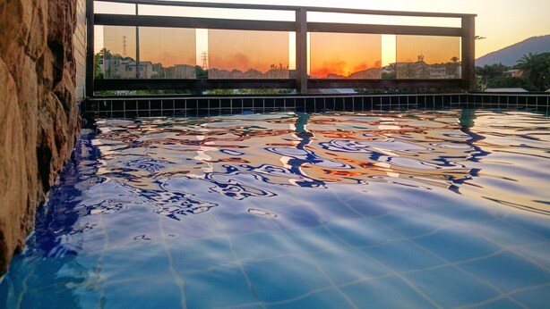 A swimming pool with a sunset view through a window, a tiled deck, and a stone wall. - Penthouse with pool, barbecue, and bathtub: Jeanne's Country House (Rio de Janeiro)