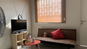 Una sala de estar con una televisión, un ventilador, una mesa roja, un sofá con un cojín rojo y una ventana con persianas.