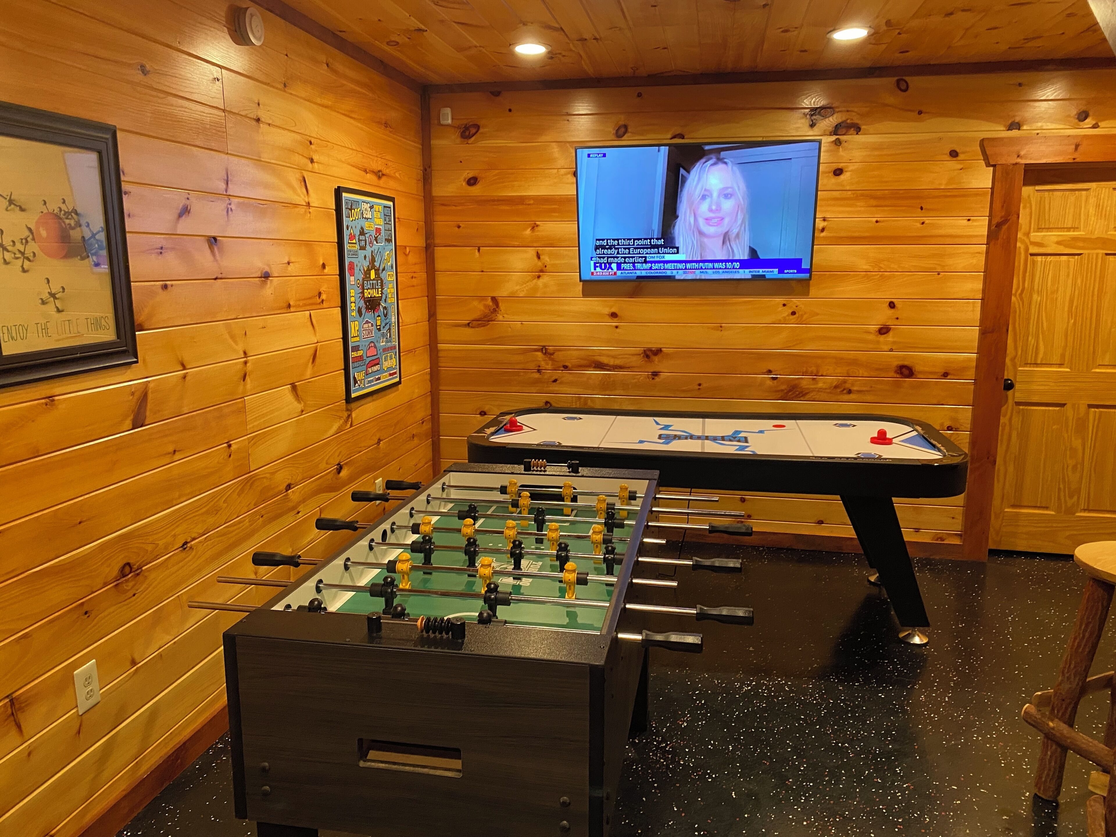 A room with wooden walls, a foosball table, a television displaying a news channel, and a framed picture on the wall.