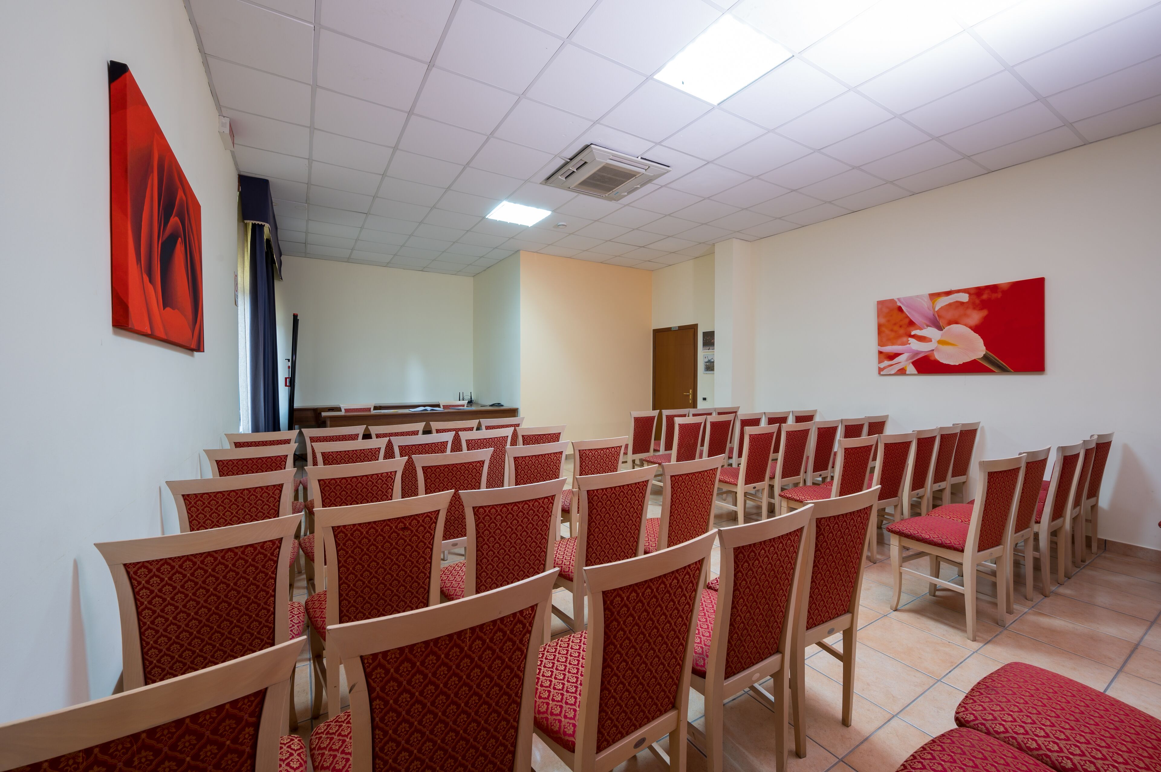 A conference room with rows of chairs and tables, a white ceiling with fluorescent lights, and two pieces of artwork on the walls.