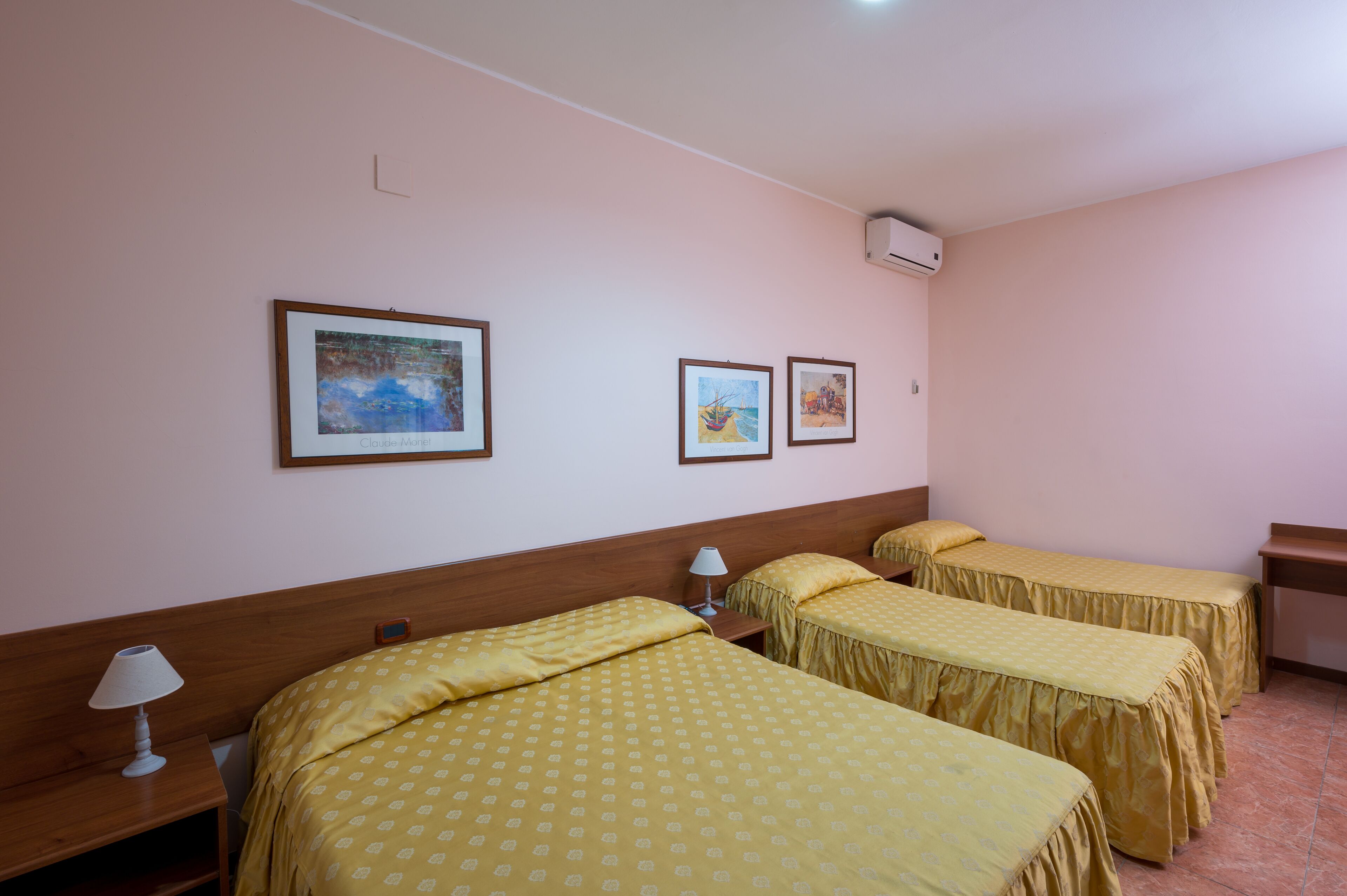 A hotel room with two beds, a wooden headboard, a nightstand with a lamp, and three framed pictures on the wall.