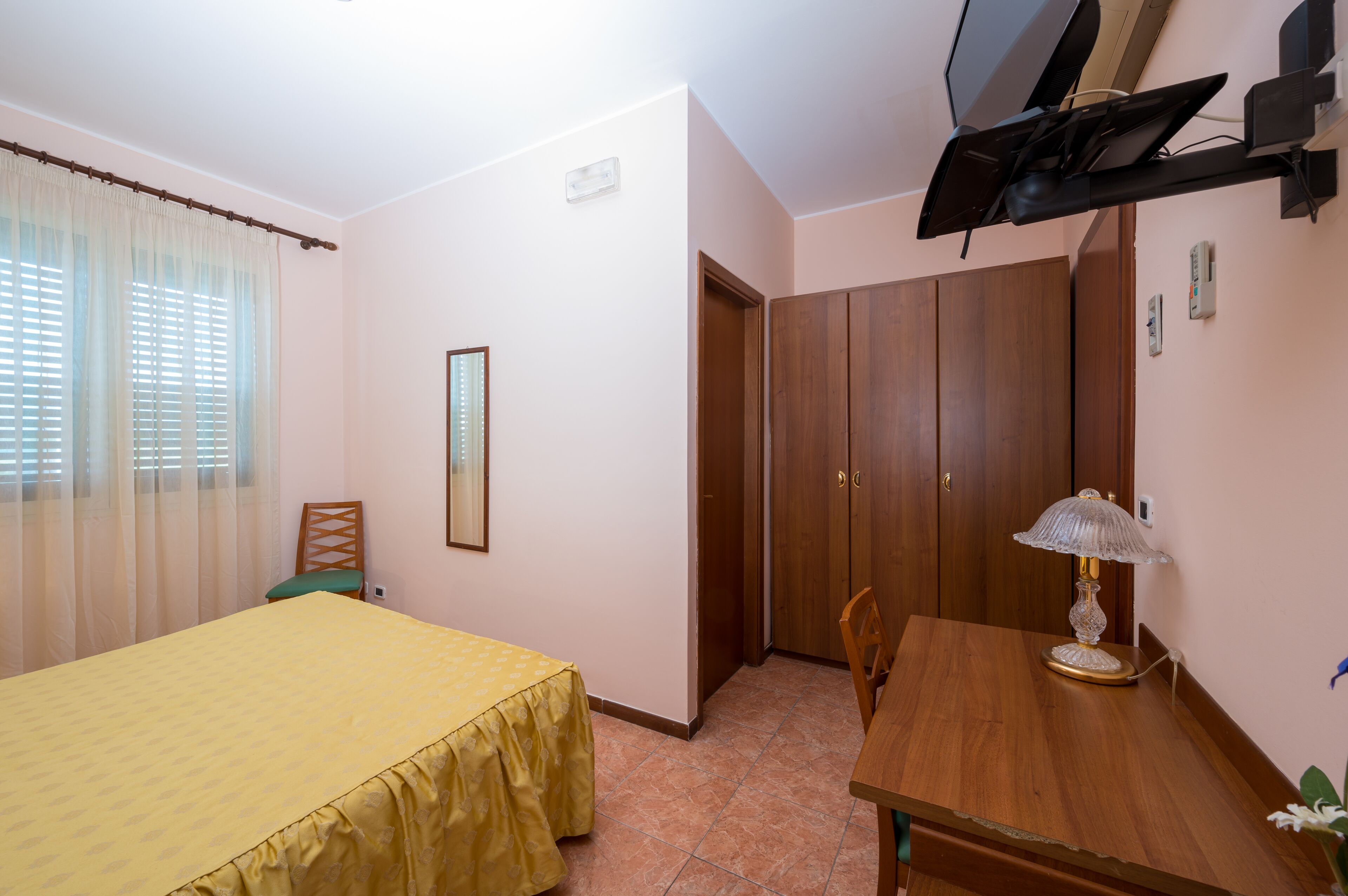 A bedroom with a bed, a wooden table, a lamp, a chair, a wardrobe, and a television.