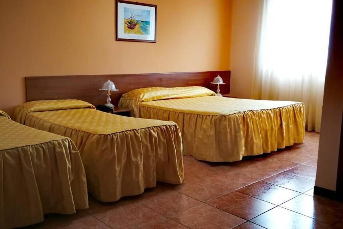 Ein Hotelzimmer mit drei Betten, jedes mit einer gelben Tagesdecke und passenden Kissen. Über den Betten hängt ein gerahmtes Bild, und auf der rechten Seite befindet sich ein Fenster mit Vorhängen.