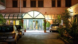 Entrada de hotel con galería techada, automóviles estacionados y macetas con plantas.