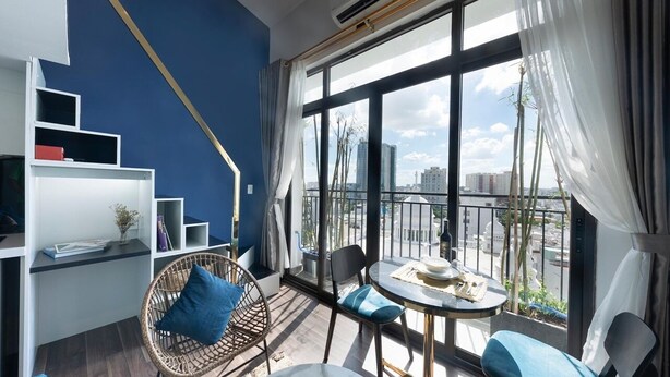 Quarto moderno com parede azul de destaque, varanda com vista para a cidade, mesa redonda e cadeiras de vime.