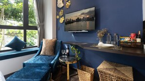 Uma sala de estar moderna com um sofá azul, uma mesinha redonda com uma garrafa de vinho, copos e uma planta. Uma televisão de tela plana exibe uma vista da cidade.