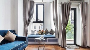 Uma sala de estar moderna com um sofá azul, uma pequena mesa redonda com um vaso, e vista para um edifício através de uma janela com cortinas.