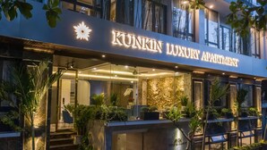 Um edifício de apartamentos de luxo chamado 'KUNKIN LUXURY APARTMENT', com design moderno, entrada de vidro e plantas ao ar livre.