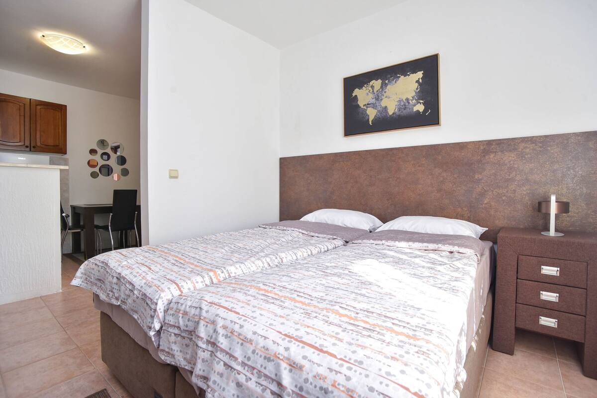 A bedroom with a bed, a nightstand, a dresser, a map on the wall, and a kitchen area in the background.
