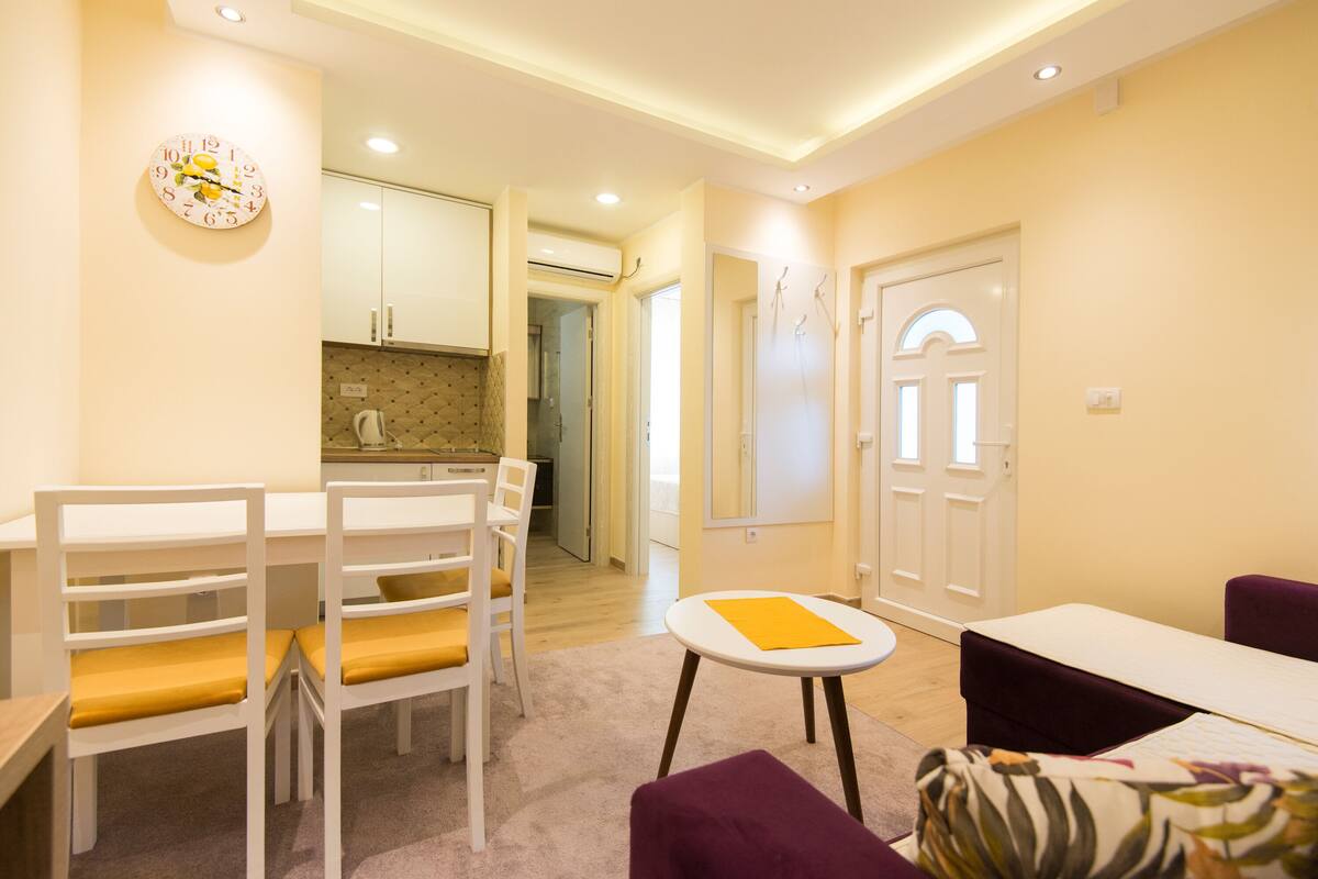 A modern living room with a dining area, a kitchen in the background, and a door leading to another room.