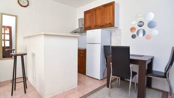 A compact kitchen with wooden cabinets, a white refrigerator, a dining table with two chairs, and a mirror on the wall.