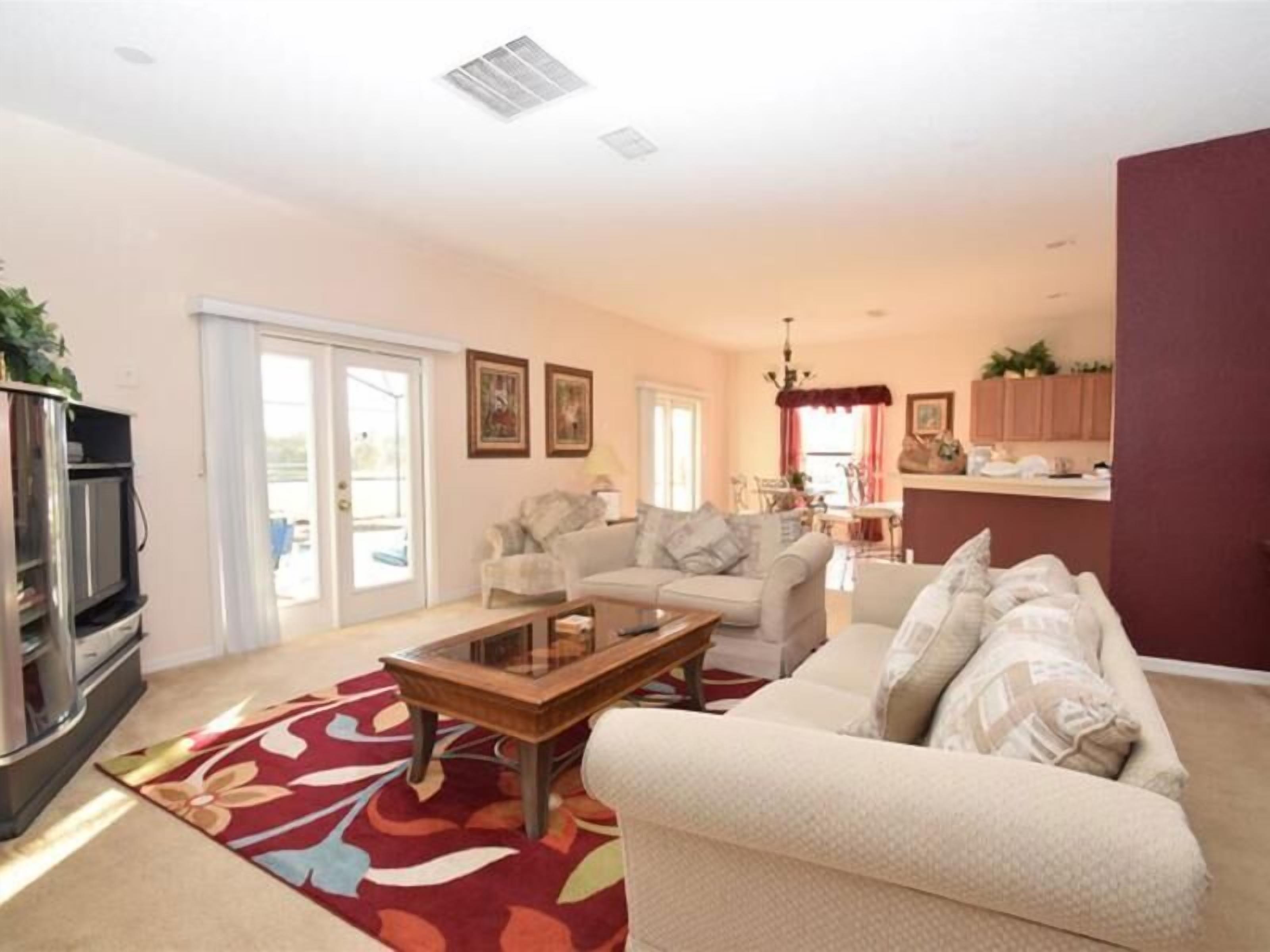 A living room with a sofa, a coffee table, and a television. There is a kitchen area in the background with cabinets and a window.