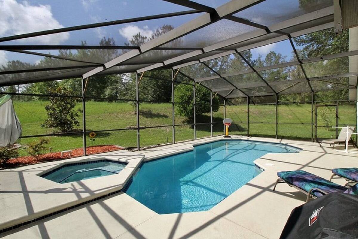 A covered outdoor swimming pool with a basketball hoop, surrounded by a screened area and a grassy landscape.