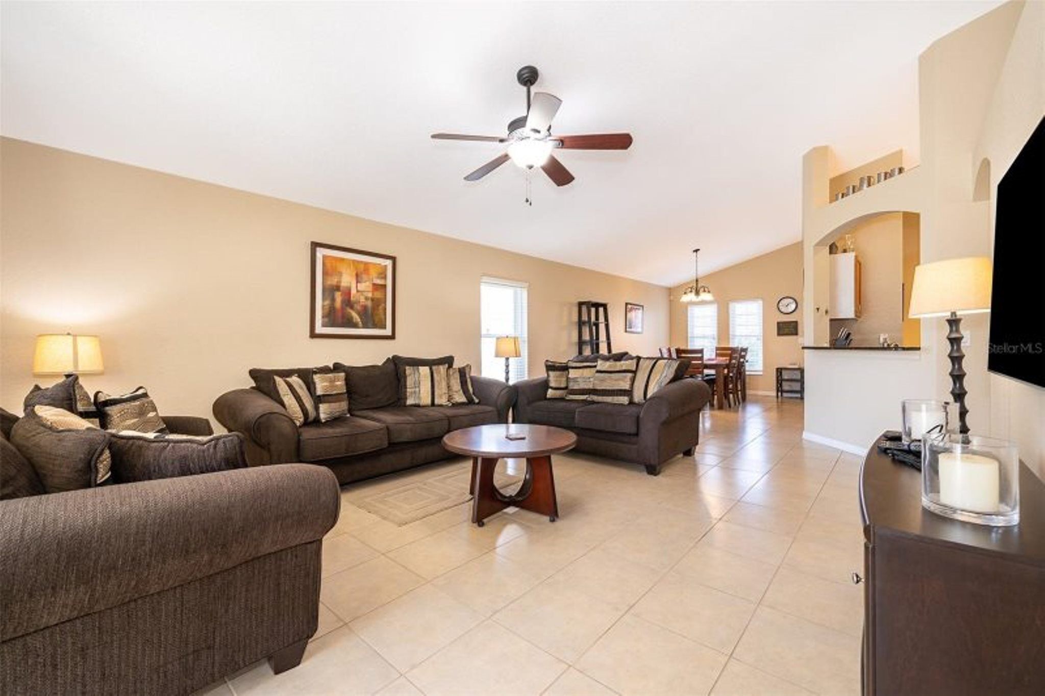 A spacious living room with two sofas, a coffee table, and a ceiling fan. A kitchen area is visible in the background.