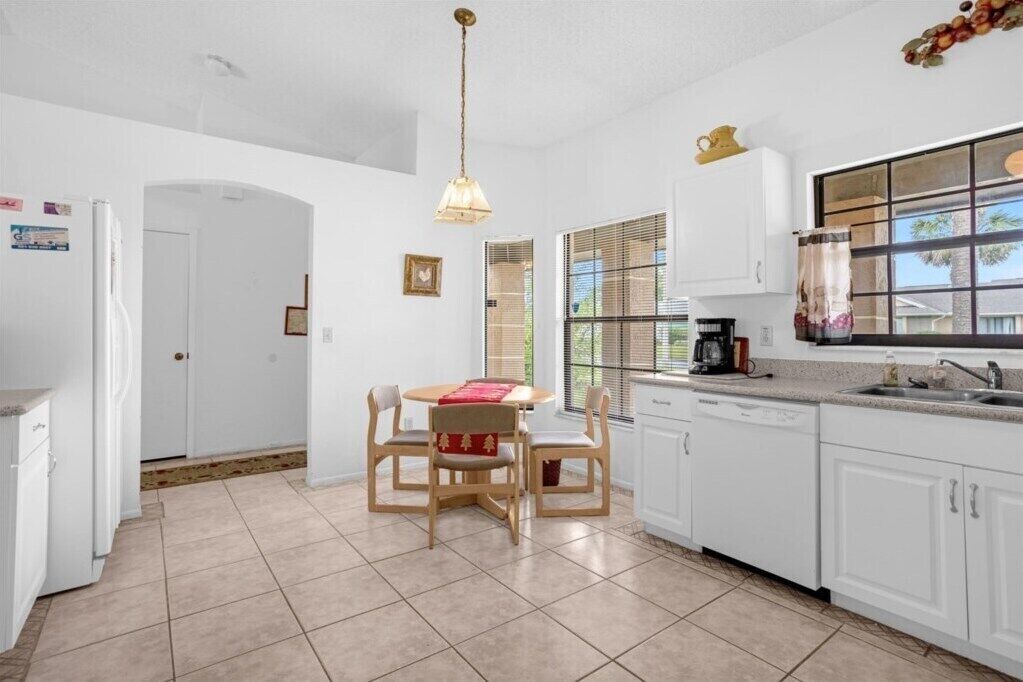 A kitchen with white cabinets, a dining table with chairs, a hanging light fixture, and a window with blinds.
