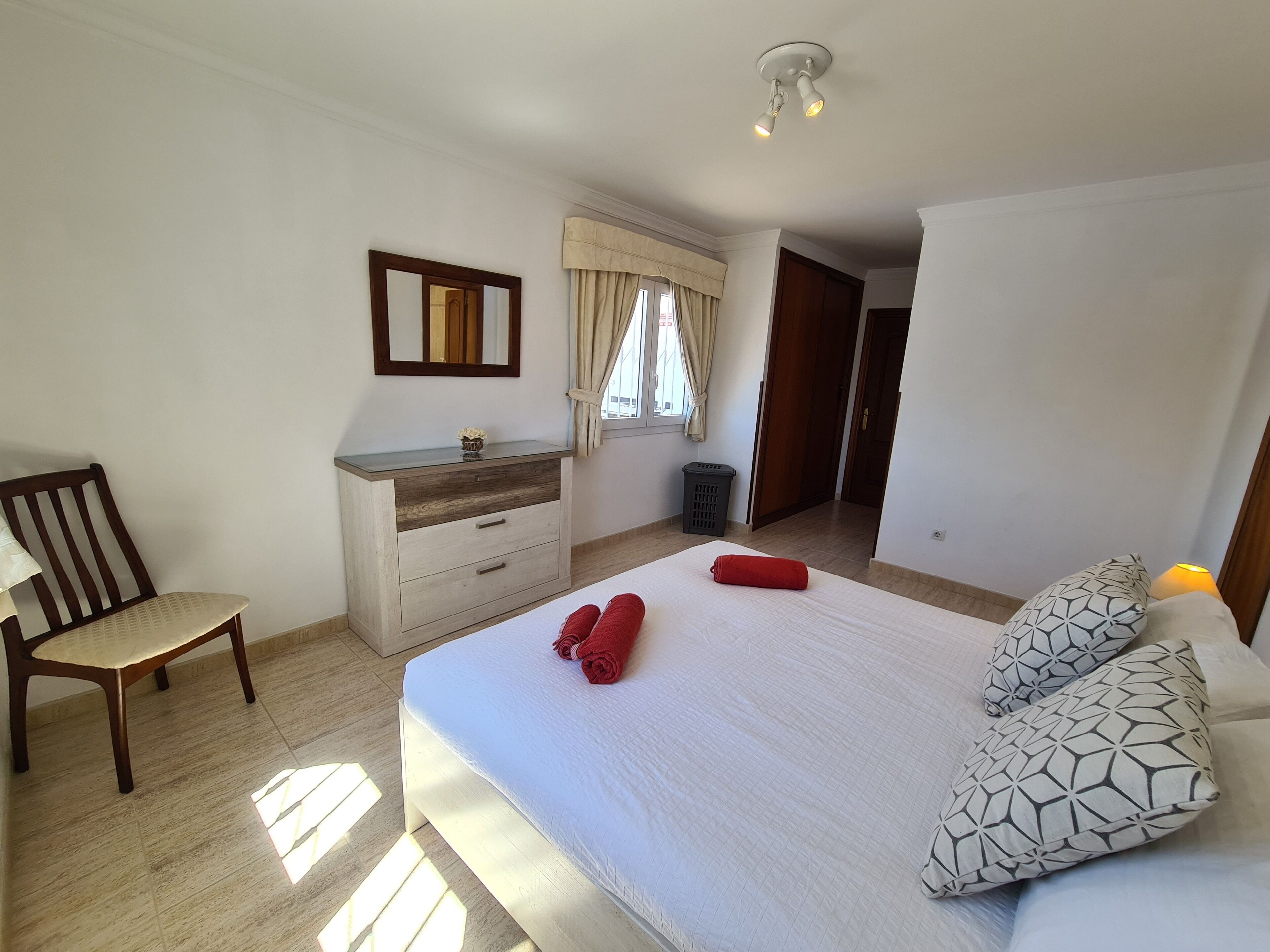 A bedroom with a bed, a wooden chair, a dresser, a mirror, and a window with curtains.