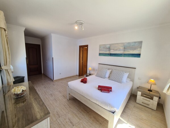 A bedroom with a bed, bedside tables, a nightstand, a wall-mounted lamp, a painting, and a wooden dresser.