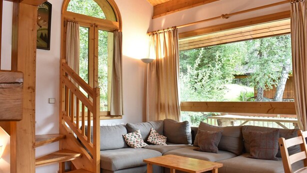 A cozy room with a wooden staircase, a sofa with cushions, a small wooden table, and large windows with curtains.