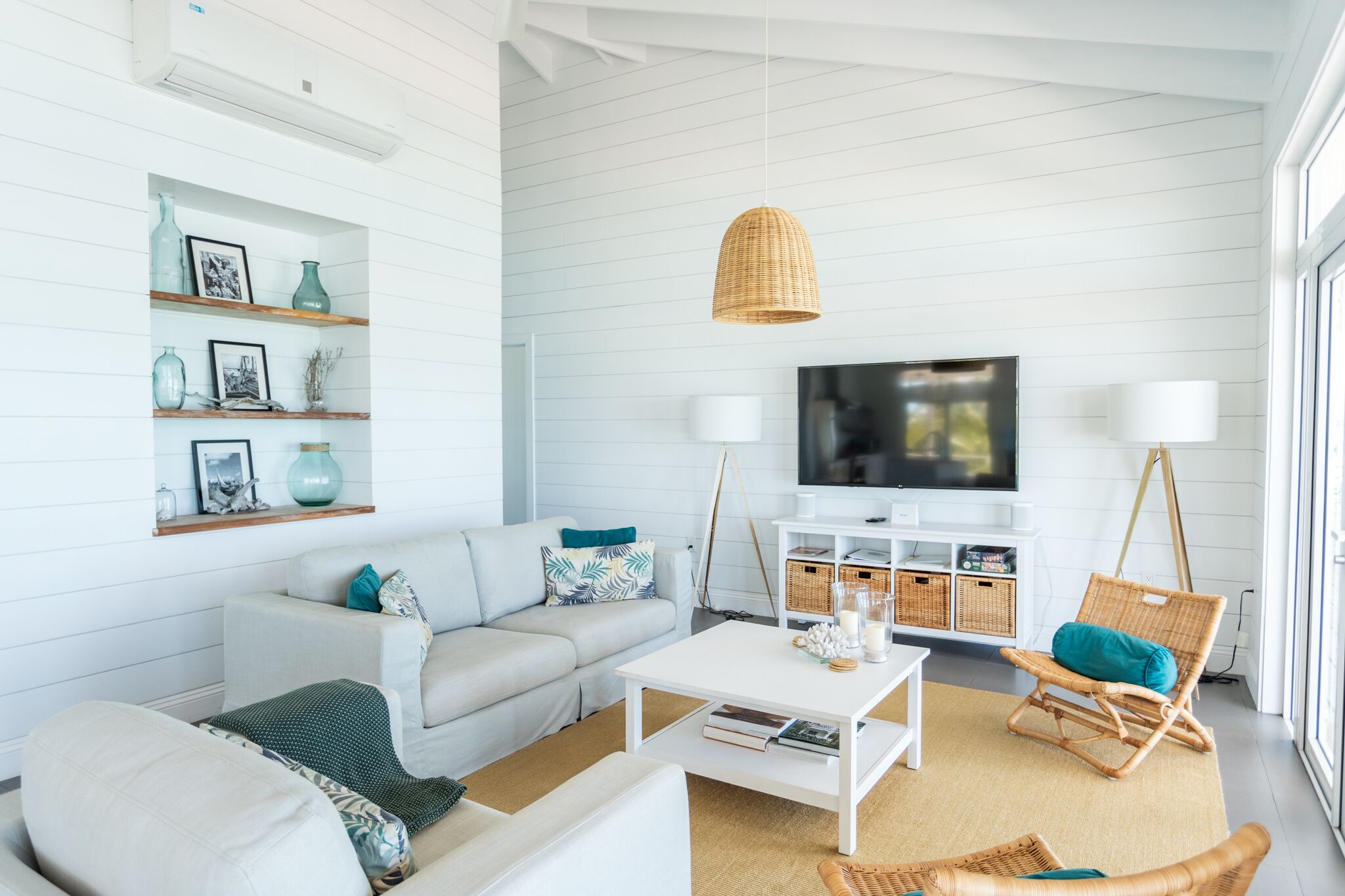 Un salon moderne avec un canapé blanc, une table basse, des fauteuils en osier, un téléviseur et des étagères ornées d’objets décoratifs.