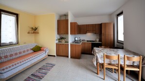 Een compacte woonruimte met een bank, een eettafel met stoelen, houten kasten, een keukenhoek en een raam met gordijn.