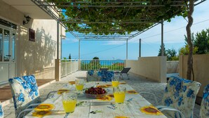 A covered outdoor dining area with a table set for two, featuring a floral tablecloth and sunflowers, a bowl of cherries, and glasses of orange juice. - PENDO 3 apartment with 4 bedrooms, sea view, close to beaches and Dubrovnik (Mlini)