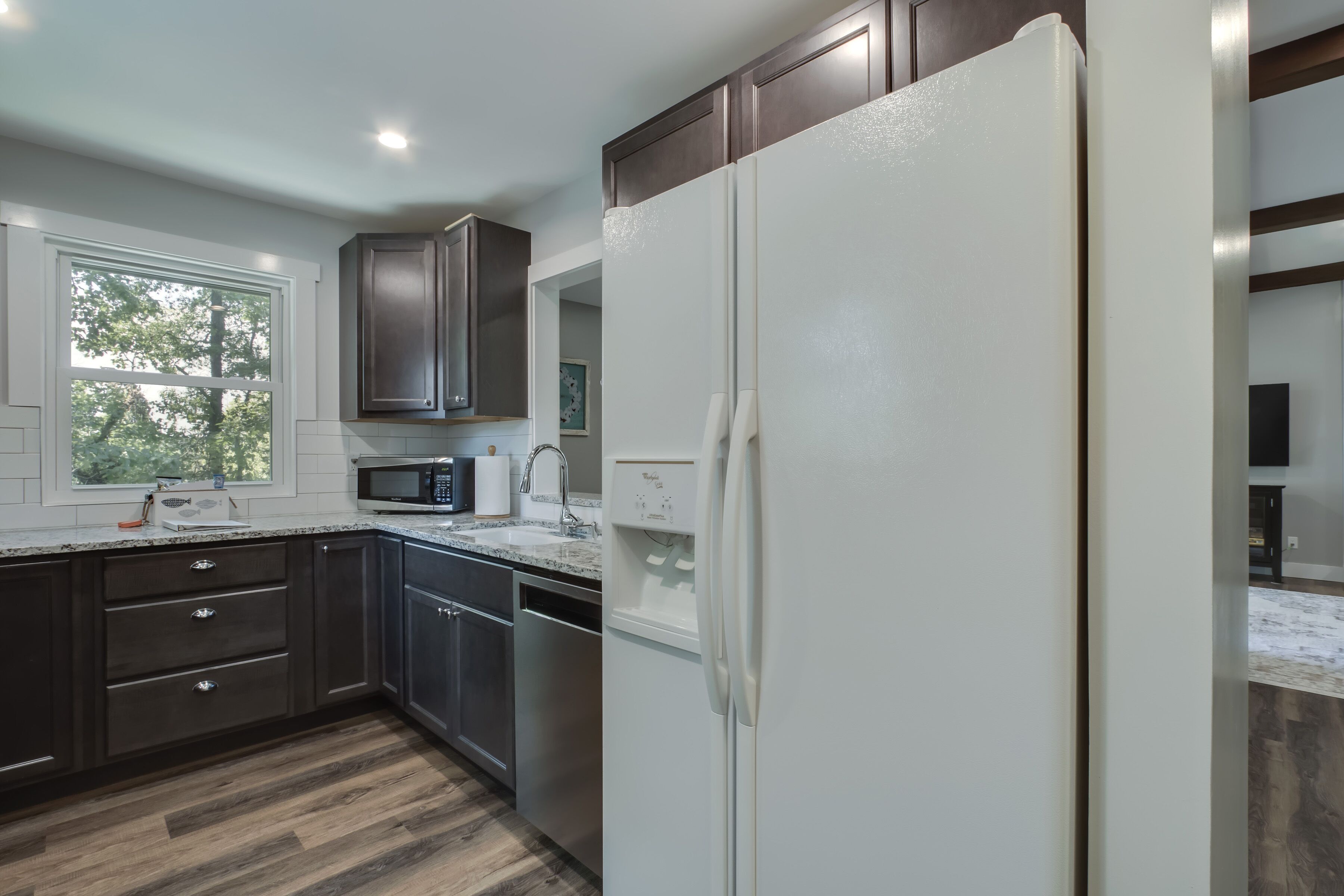 A modern kitchen with dark cabinets, a white refrigerator, stainless steel appliances, and a window with a view of trees.