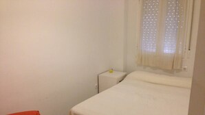 Una habitación pequeña y sencilla con una cama individual, una mesita de noche, una silla y una ventana con cortinas ligeras.