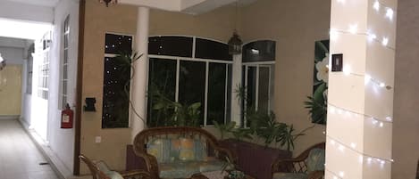 A living room with wicker furniture, a patterned sofa, and a small table with books. There are potted plants and a window with curtains.