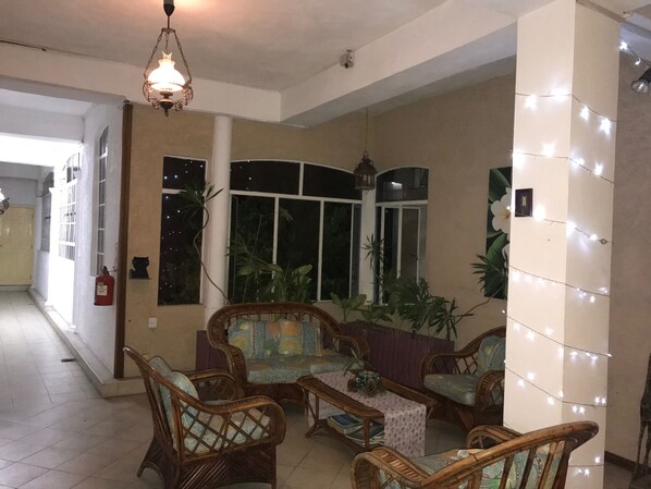 Sala de estar con muebles de mimbre, un sofá estampado y una mesita con libros. Hay plantas en macetas y una ventana con cortinas.