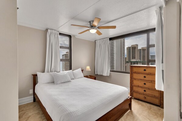 Un dormitorio con cama, cómoda, mesita de noche, ventilador de techo y ventanas grandes con cortinas.