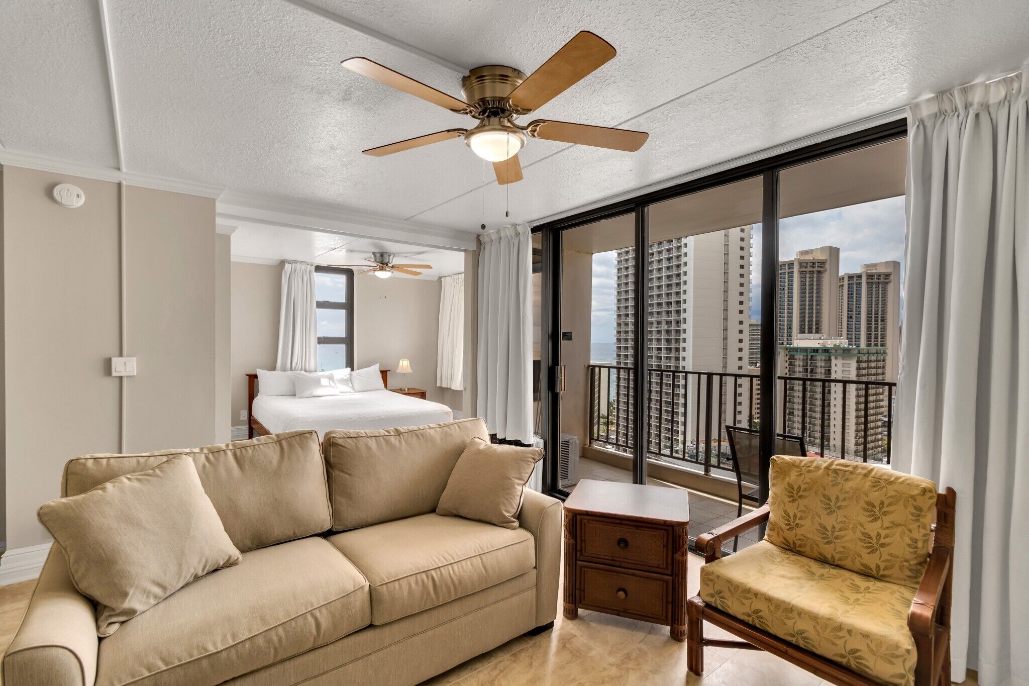 A hotel room with a large bed, a sofa, a chair, a small table, a ceiling fan, and a balcony with a city view.