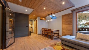 Un interior moderno con techo de madera, una zona de comedor con sillas y una mesa, un sofá y un ventanal con vistas a casas y árboles.