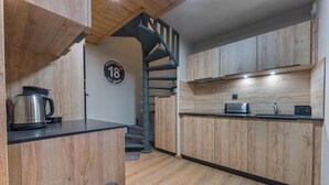Una cocina compacta con armarios de madera, un hervidor de acero inoxidable, un tostador y una encimera negra. Se ve una escalera de caracol al fondo.