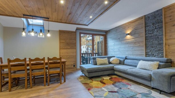 Una sala de estar acogedora con techo de madera, un sofá seccional gris, una mesa de comedor de madera con sillas y una alfombra colorida.