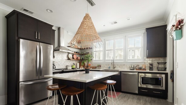 Cocina moderna con armarios oscuros, electrodomésticos de acero inoxidable, una isla central con taburetes y una lámpara colgante.
