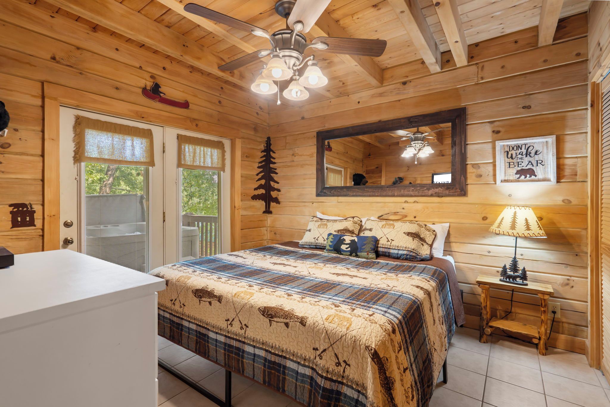 Un dormitorio estilo cabaña con cama, escritorio, espejo, ventilador de techo y ventana con vista a árboles.