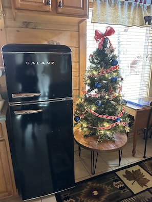 Une cuisine avec un réfrigérateur noir, un sapin de Noël et une fenêtre avec des stores.
