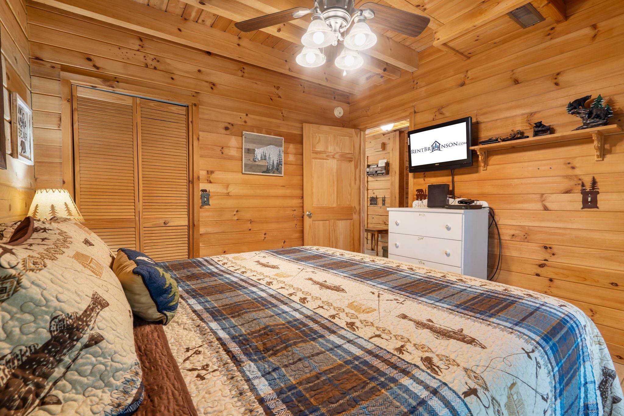 A wooden cabin bedroom with a bed, a dresser, a television, and a ceiling fan.