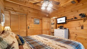 A wooden cabin bedroom with a bed, a dresser, a television, and a ceiling fan.
