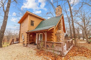 Une cabane en rondins avec un porche, une cheminée et une brouette à l’extérieur.