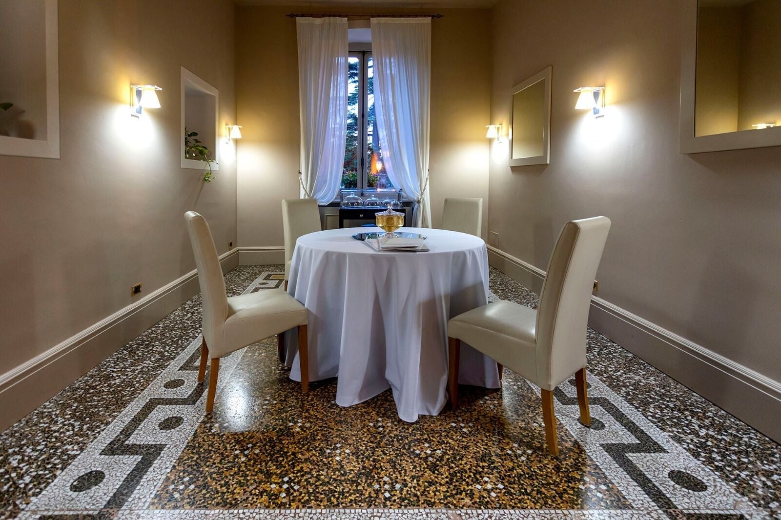 A dining area with a round table, four chairs, a patterned floor, and a view through a window with curtains.