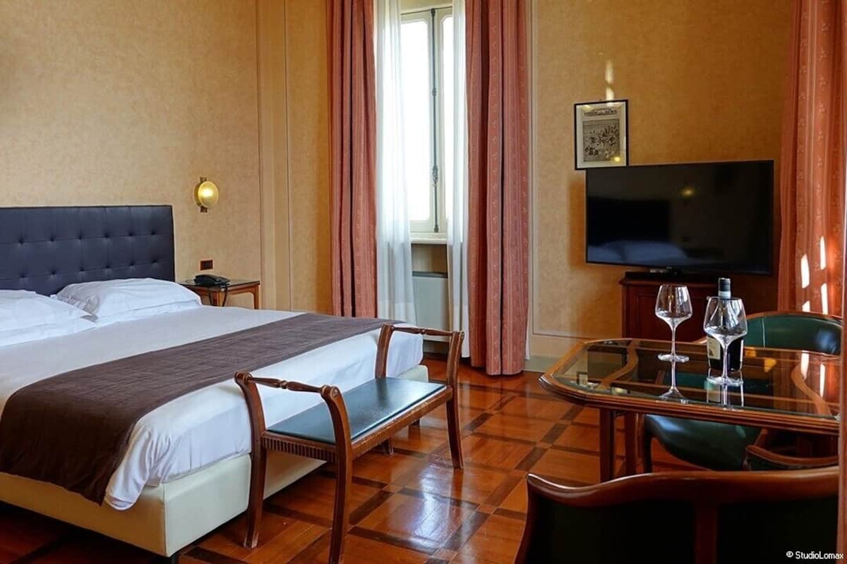 Hotellihuone, jossa on suuri sänky, televisio, lasinen pöytä viinilaseineen ja tuoli.