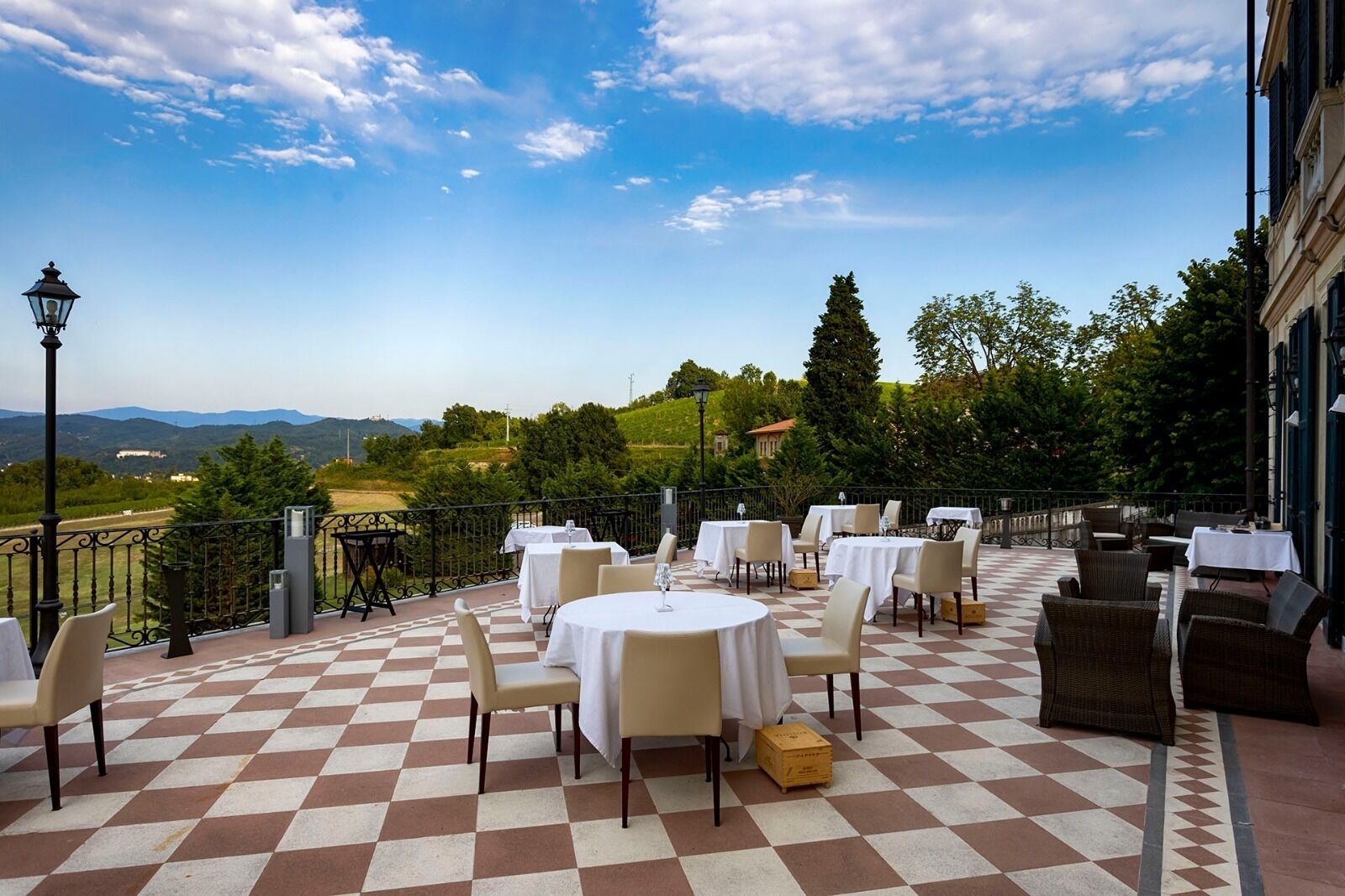 An outdoor patio with checkered flooring, round tables, and chairs, overlooking a scenic landscape with trees and distant hills.