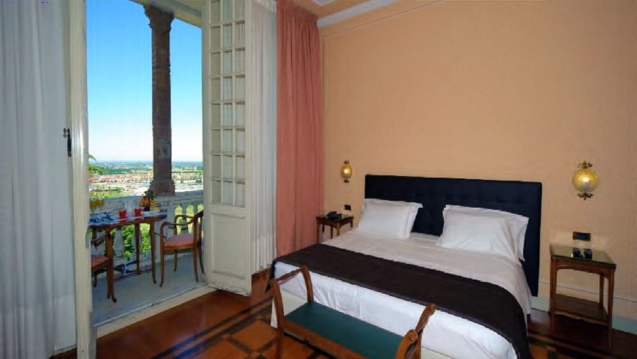Habitació d'hotel amb un llit gran, un balcó amb taula i cadires, i vista a la ciutat.