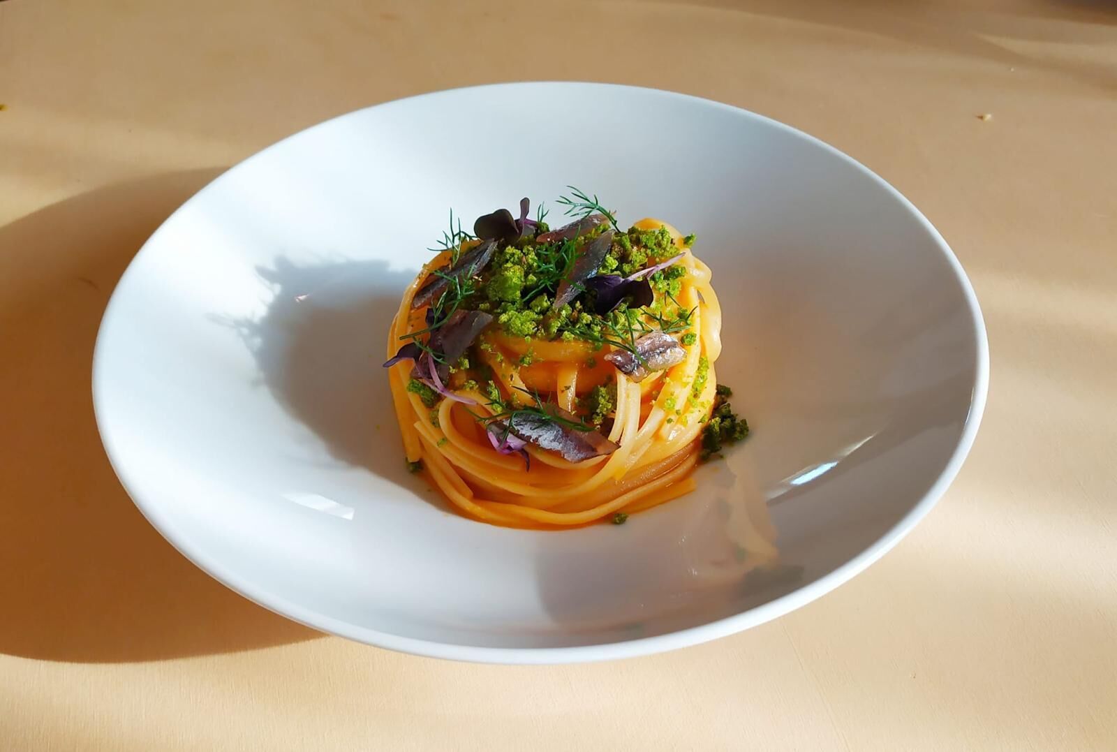 Un piatto bianco con un primo a base di pasta, guarnito con erbe verdi e foglie viola, su una superficie beige.