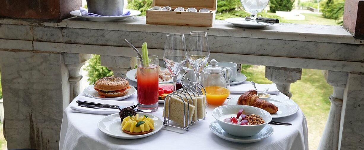 A table set with breakfast items including a croissant, a sandwich, a fruit bowl, a glass of juice, and a drink with a straw.