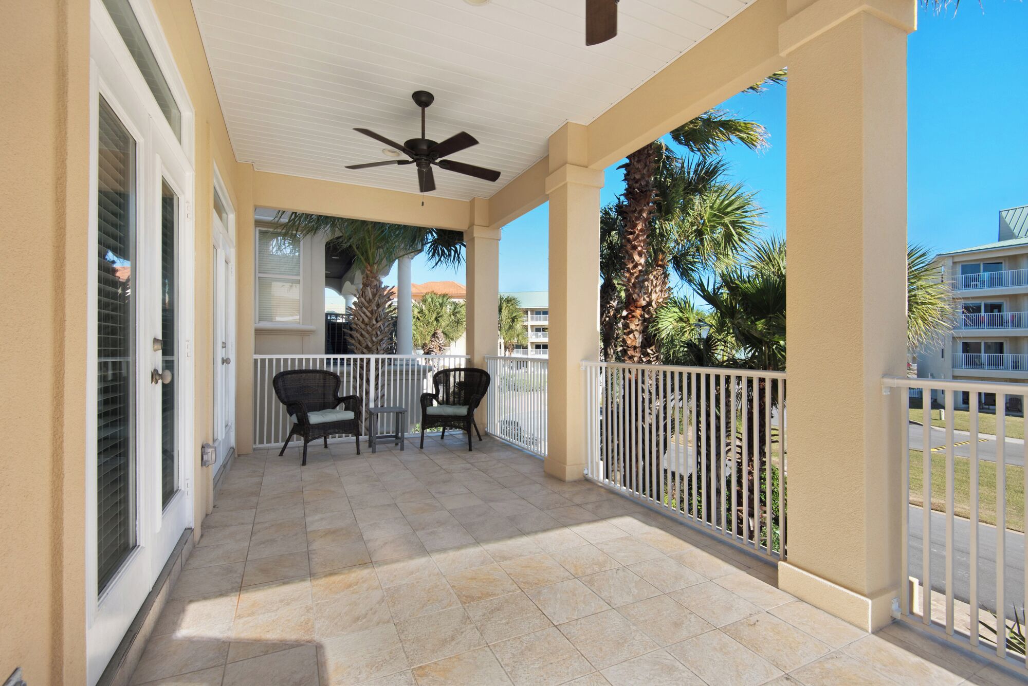 A covered outdoor area with two chairs and a table, a ceiling fan, and a view of palm trees and buildings.