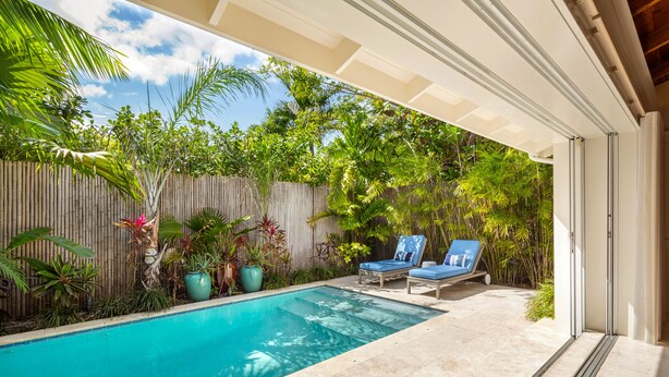 Une piscine entourée d’une clôture en bois et agrémentée de plantes en pot, deux chaises longues bleues et un patio couvert.